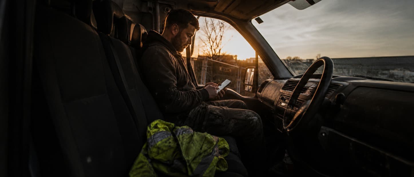 Tradesman in workwear in his van at sunset, sending a quote on his phone, folded high-vis on the seat.